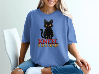 Black cat with yellow eyes sits on a blue t-shirt with "KNEEL BEFORE ME" text.