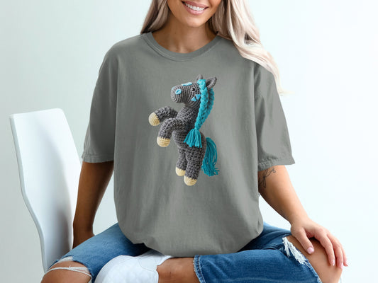 Crocheted grey horse with blue mane and tail on a grey t-shirt.