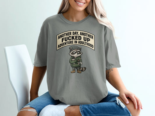 Graphic t-shirt featuring a raccoon with "Another day, another fucked up adventure in adulthood" text.