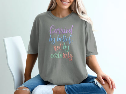 Gray t-shirt with "Carried by belief, not by certainty" in colorful gradient text.