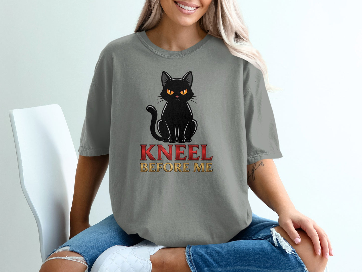 Woman wearing a grey t-shirt with a black cat and "KNEEL BEFORE ME" text.