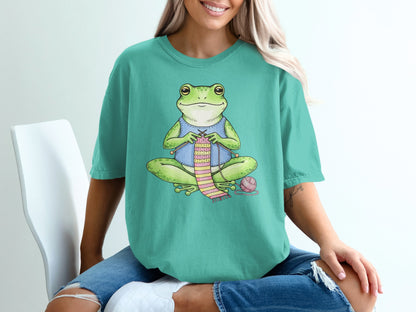 Frog knitting a pink and green striped scarf with yarn and needles.