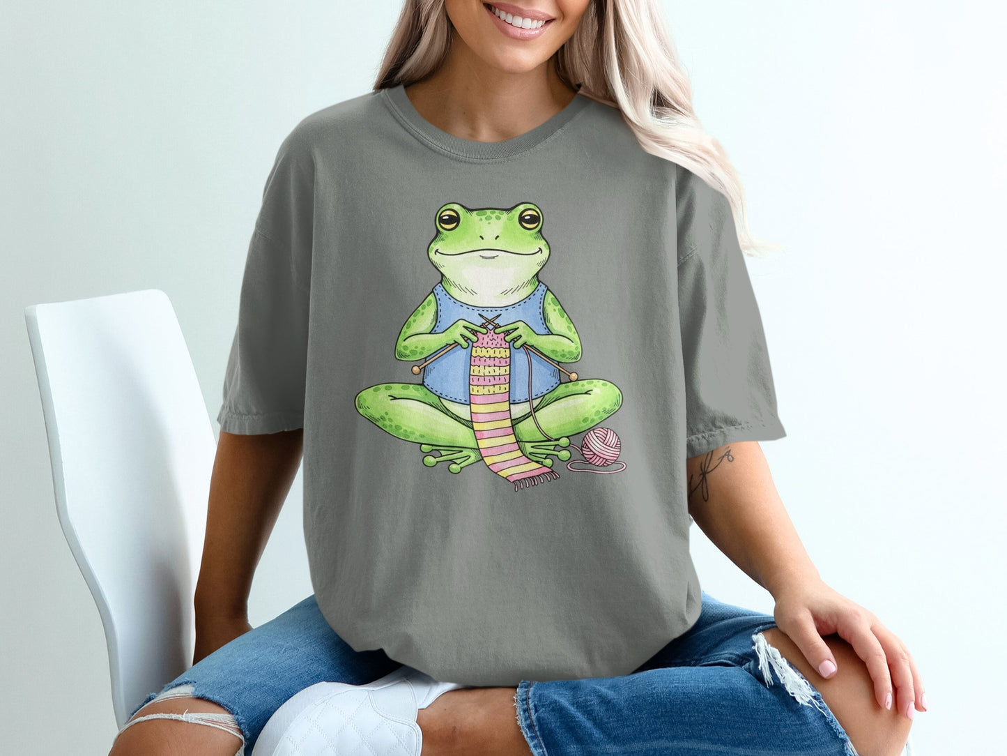 Cartoon frog knitting a pink and green scarf with yarn and needles.