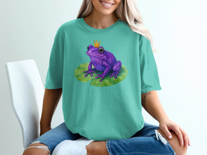 Purple frog wearing a crown on a lily pad, on a teal t-shirt.