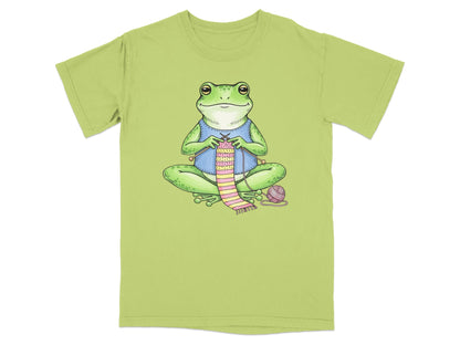 Green t-shirt with illustration of a frog knitting a pink and green striped scarf.