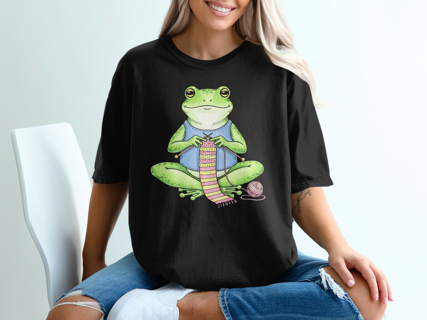 Frog wearing blue shirt knitting a pink scarf on a black t-shirt.