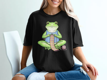 Frog wearing blue shirt knitting a pink scarf on a black t-shirt.