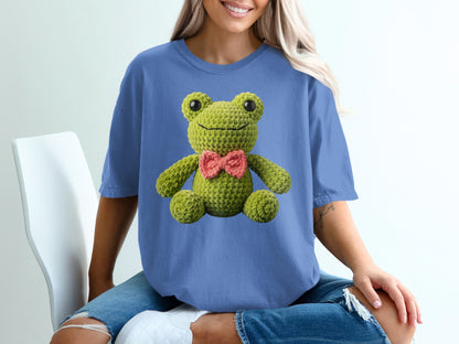Crocheted green frog with a pink bow tie on a blue t-shirt.