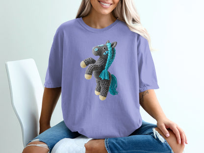 Crocheted gray unicorn with blue mane and tail on lavender t-shirt.