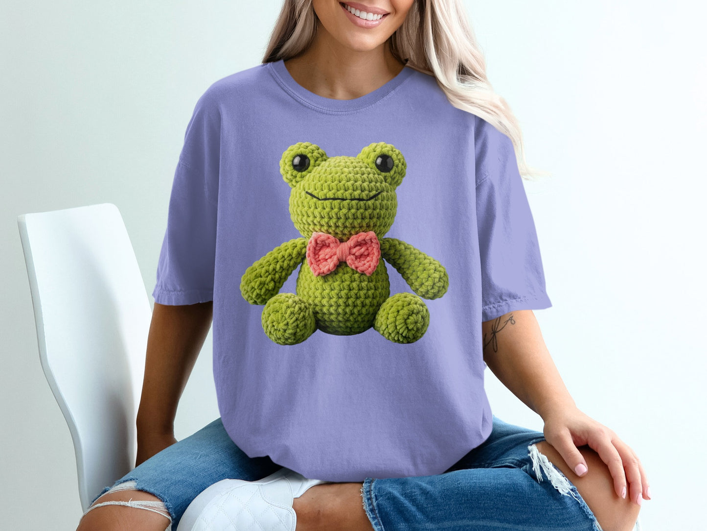 Crocheted green frog with pink bow tie on purple t-shirt.