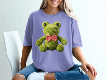 Crocheted green frog with pink bow tie on purple t-shirt.