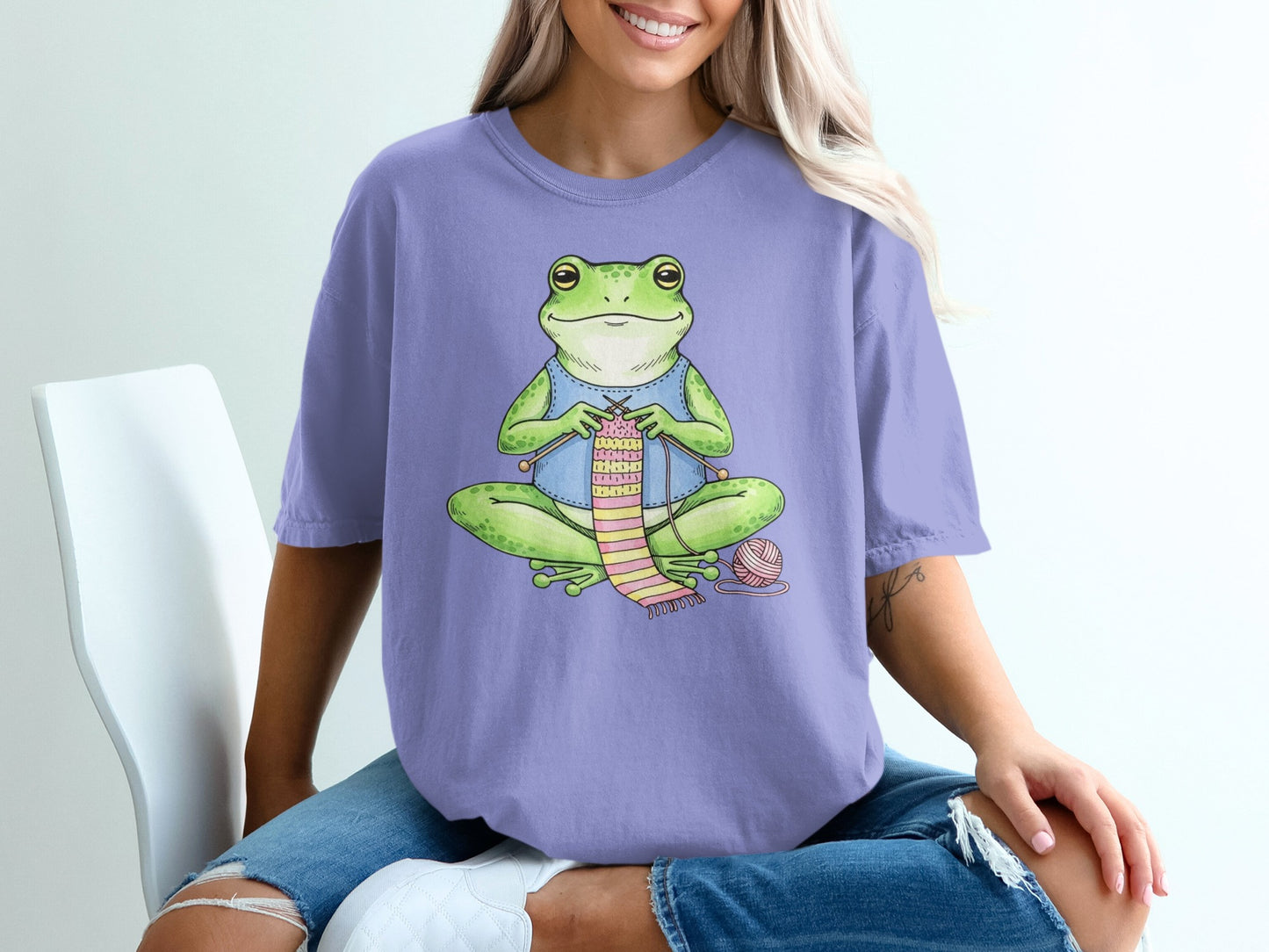 Frog wearing blue overalls knitting pink striped sweater with yarn.