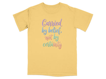 Yellow t-shirt with "Carried by belief, not by certainty" in colorful cursive text.