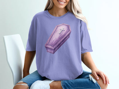 Purple t-shirt featuring a watercolor illustration of a coffin with a cross.